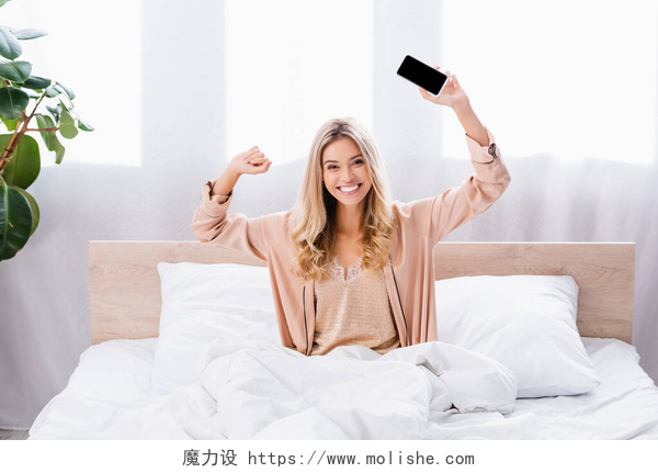 早晨从床铺上醒来的女人拿着手机早上穿着睡衣拿着智能手机躺在床上的快乐女人 